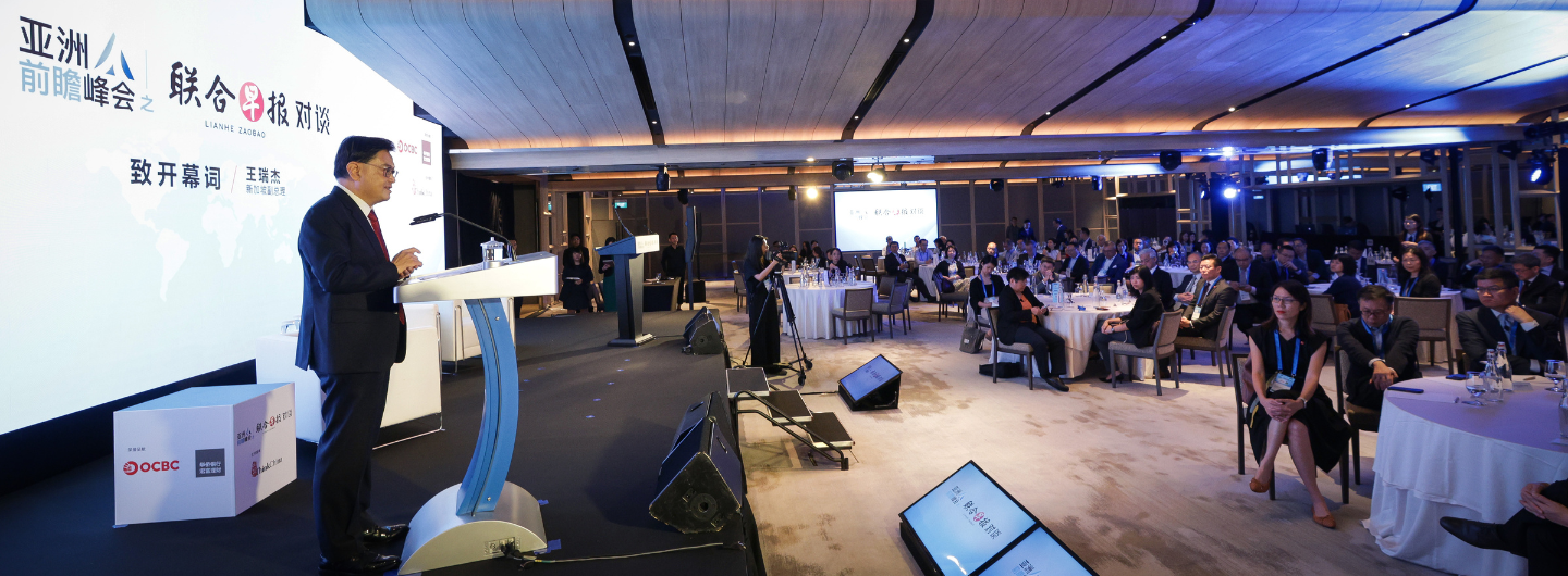 Man in suit speaks at podium with OCBC logo, to audience seated at round tables.